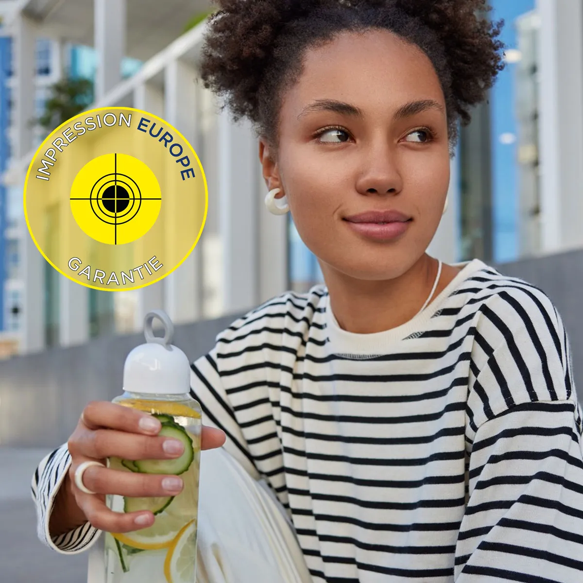 Jeune femme souriante tenant une gourde infusée de citron et concombre, portant un haut marinière, avec le logo 'Impression Europe Garantie' en arrière-plan